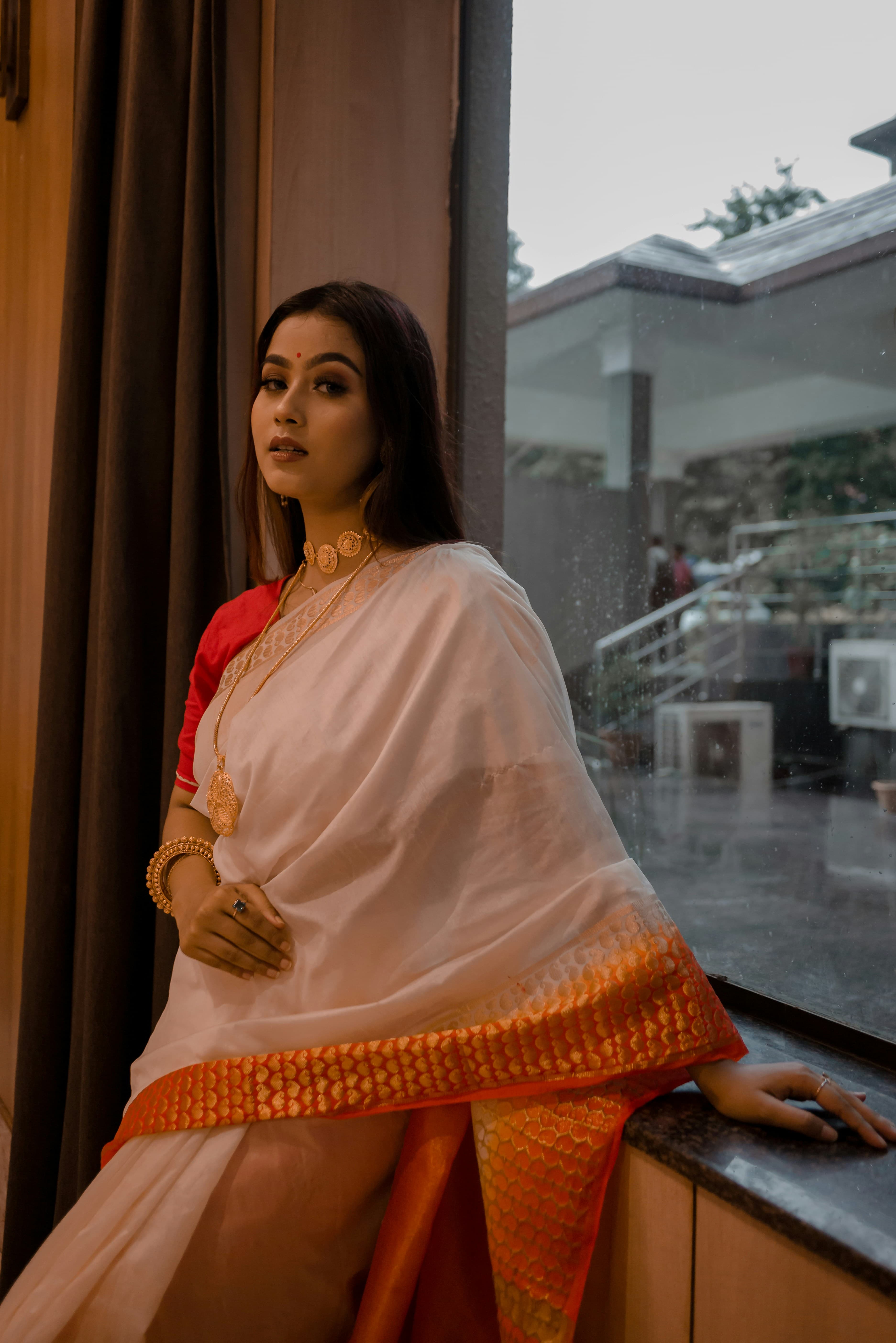 Woman in a white saree with an orange zari border, seated by a window